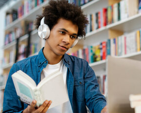 Ein junger Mann mit Kopfhörern liest ein Buch, während er an einem Laptop in einer Bibliothek sitzt, umgeben von Regalen voller Bücher.