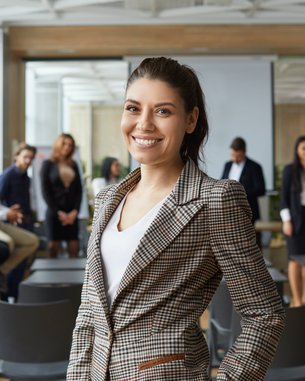 A smiling woman in a checked blazer stands in the foreground of a modern conference room, while several people are talking to each other in the background.