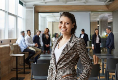 A smiling woman in a checked blazer stands in the foreground of a modern conference room, while several people are talking to each other in the background.