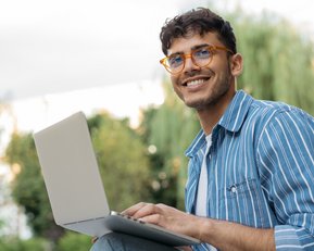 Junger Mann mit Brille sitzt im Freien und arbeitet an einem Laptop, lächelnd und entspannt.
