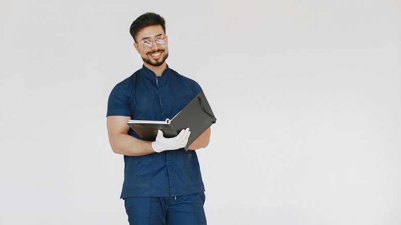 Ein lächelnder Mann in blauer Arbeitskleidung hält einen Ordner in der Hand. Er trägt eine Brille und weiße Handschuhe, während er vor einem neutralen Hintergrund steht.