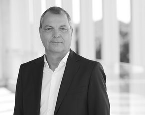 Ein Mann in einem schwarzen Anzug mit weißem Hemd steht vor einer hellen, modernen Architektur.