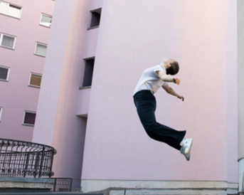 Eine Person in formeller Kleidung springt in die Luft, mit einem Ausdruck von Bewegung und Energie. Im Hintergrund sind pinkfarbene Wände eines Gebäudes sichtbar, die eine moderne architektonische Umgebung schaffen.