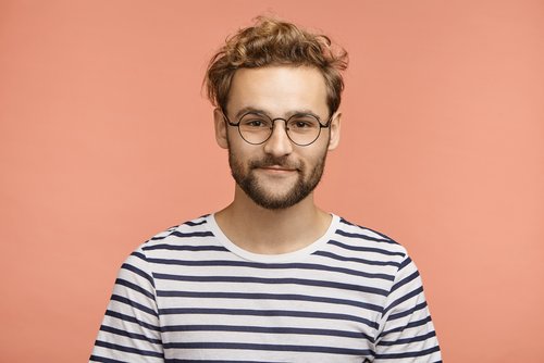 Junger Mann mit lockigem, blondem Haar und Brille, trägt ein gestreiftes T-Shirt vor einem einfarbigen, rosa Hintergrund.