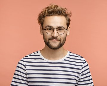 Junger Mann mit lockigem, blondem Haar und Brille, trägt ein gestreiftes T-Shirt vor einem einfarbigen, rosa Hintergrund.