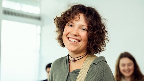 Lächelnde junge Frau mit lockigem Haar, die eine Gitarre hält. Im Hintergrund sind weitere Personen unscharf zu sehen.