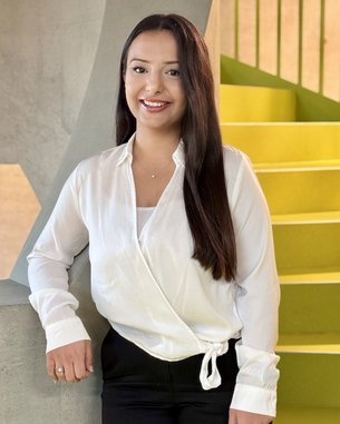 Eine Person mit langen, braunen Haaren trägt eine weiße Bluse und schwarze Hose. Sie steht an einer Wand und lächelt. Im Hintergrund sind gelbe Treppenstufen sichtbar.