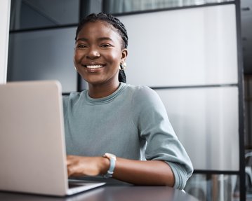Eine lächelnde Frau mit Zopf sitzt an einem Tisch und arbeitet an einem Laptop in einem modernen Büro.