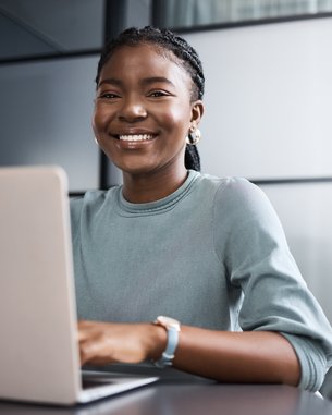 A smiling woman with a braid sits at a table working on a laptop in a modern office.