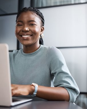 A smiling woman with a braid sits at a table working on a laptop in a modern office.