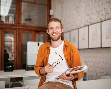 Student mit Brille und Unterlagen in der Hand lacht in die Kamera