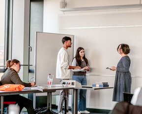 Drei Personen stehen in einem Klassenzimmer und diskutieren. Eine Person hält Notizen, während eine andere spricht. Im Hintergrund sind Schüler an Tischen mit Wasserflaschen und Materialien zu sehen.