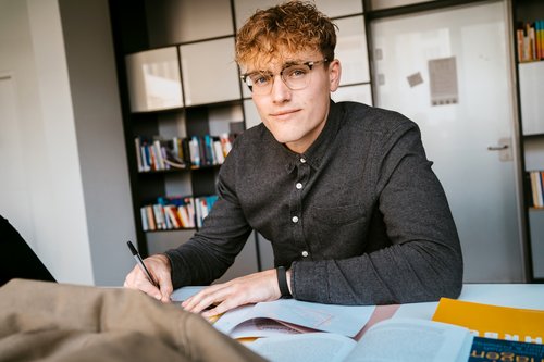 A young man with curly hair and glasses is sitting at a table, writing in a notebook, with books in front of him.