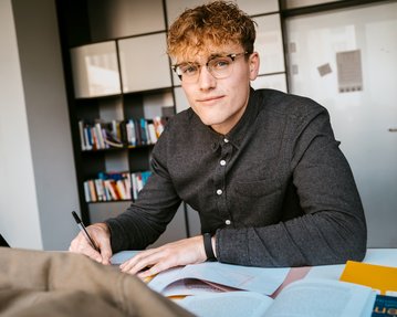 A young man with curly hair and glasses is sitting at a table, writing in a notebook, with books in front of him.