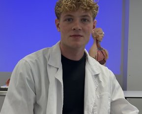 Ein junger Mann in einem weißen Laborkittel sitzt in einem Klassenzimmer mit einem anatomischen Modell im Hintergrund.