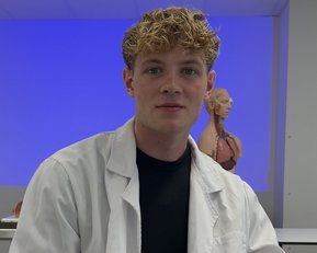 Ein junger Mann in einem weißen Laborkittel sitzt in einem Klassenzimmer mit einem anatomischen Modell im Hintergrund.