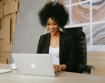 A woman with curly hair is sitting at a desk, working on a laptop. She is wearing a black blazer and glasses, smiling as she looks at the screen.