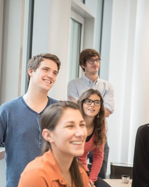 A group of five young adults is smiling and attentively looking in a bright room with large windows. They appear to be participating in a discussion or presentation.