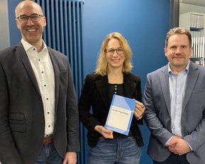 Publikationspreis Gesundheitspsychologie Drei Personen stehen in einem modernen Büro. Eine Person hält ein Buch in der Hand, während die anderen lächeln und in die Kamera schauen.