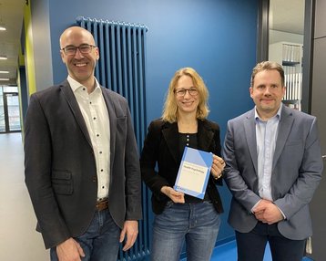 Drei Personen stehen in einem modernen Büro. Eine Person hält ein Buch in der Hand, während die anderen lächeln und in die Kamera schauen.