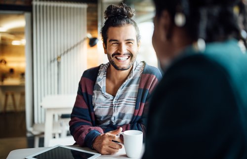 Ein Mann mit lockigem Haar und einem gestreiften Pullover sitzt an einem Tisch, lächelt und hält eine Tasse in der Hand.