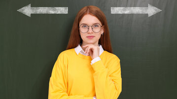 Eine junge Frau mit langen, roten Haaren und Brille trägt einen gelben Pullover und steht vor einer grünen Tafel mit Pfeilen nach links und rechts.