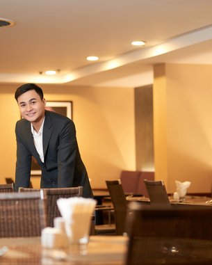 A man in a suit stands at a table in a restaurant. The surroundings are modern and well-lit. In the background, there is a person wearing a traditional hat.