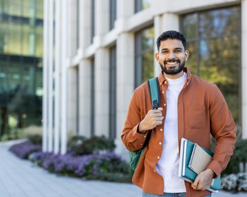 A young man with a beard is wearing an orange jacket and carrying a backpack. He is holding books and standing in front of a modern building.