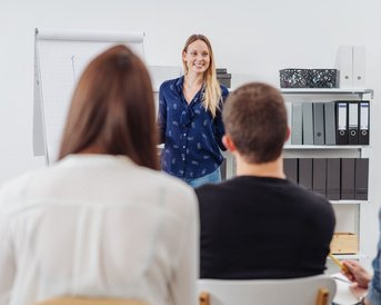 Junge, blonde Frau hält einen Vortrag vor ihren Kolleg:innen.