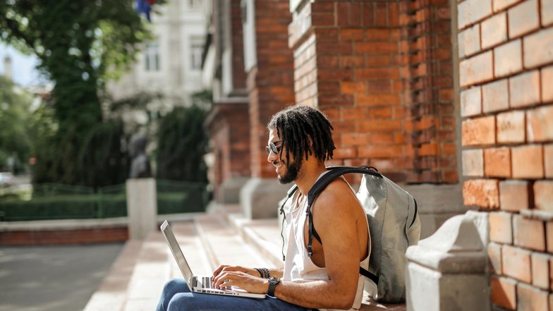 A man with dreadlocks is sitting on a staircase, working on a laptop. He’s wearing a backpack and dressed in casual attire. In the background, there are trees and a brick wall visible.
