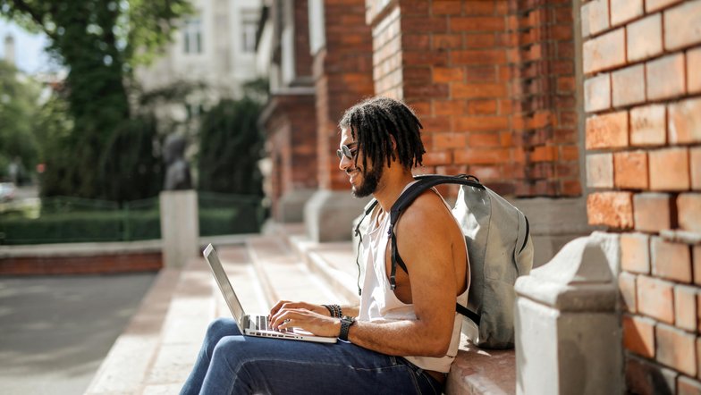 Ein Mann mit Dreadlocks sitzt auf einer Treppe und arbeitet an einem Laptop. Er trägt einen Rucksack und hat ein lässiges Outfit an. Im Hintergrund sind Bäume und eine Ziegelmauer sichtbar.