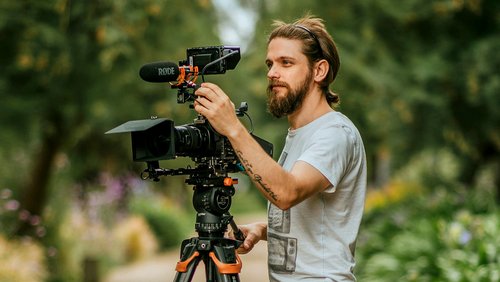 Ein Mann mit Bart stellt eine Kamera auf einem Stativ ein, während er in einem Garten steht. Die Kamera hat ein Mikrofon montiert.