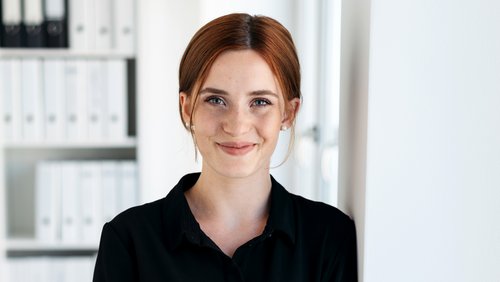 Junge Frau mit roten Haaren, die in einem Büro steht und lächelt. Im Hintergrund sind Aktenordner sichtbar.
