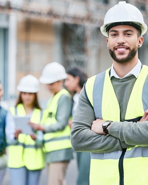 A construction manager in safety gear and a helmet stands smiling in the foreground, while a team of construction workers discusses in the background.