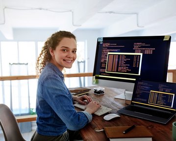 A young woman sits at a desk, working on two computers while smiling. Programming code is visible on the screens.