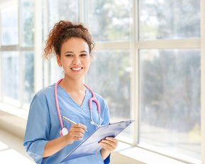 Eine junge Frau in blauer medizinischer Kleidung hält ein Clipboard und einen Stift. Sie trägt ein Stethoskop um den Hals und lächelt freundlich. Helle Fenster im Hintergrund lassen viel Licht herein.