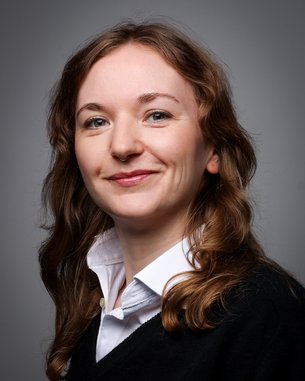 A young woman with curly brown hair and a nose piercing is wearing a black top and a white shirt.
