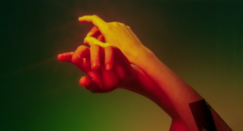 Hands in various skin tones touching and intertwining. The hands are set against a soft, multicolored background that includes shades of green and yellow.