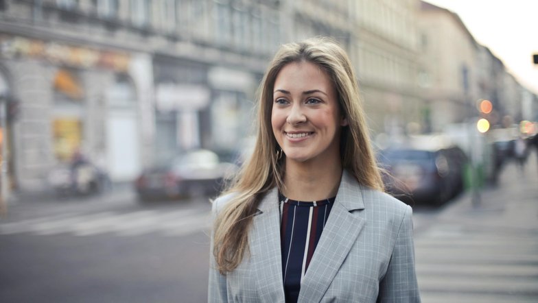 Eine lächelnde Frau mit langen, lockigen Haaren trägt einen grauen Anzug und steht auf einer Straße, umgeben von Gebäuden und Autos im Hintergrund.