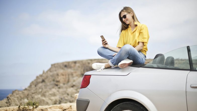 Eine Frau sitzt auf dem Kofferraum eines silbernen Autos und schaut auf ihr Smartphone. Sie trägt ein gelbes T-Shirt und Jeans, mit einer Küstenlandschaft im Hintergrund.
