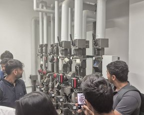 A group of people stands in a room in front of a series of technical devices with pipes and measuring instruments. Some individuals are holding smartphones in their hands.