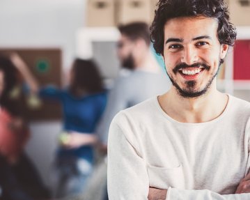 Ein junger Mann mit einem Bart lächelt und steht mit verschränkten Armen im Vordergrund. Im Hintergrund sind mehrere Personen zu sehen, die an einer kreativen Aktivität teilnehmen. Die Umgebung wirkt modern und einladend.