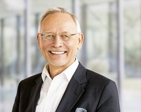 Ein lächelnder Mann mit Brille trägt einen schwarzen Anzug und ein weißes Hemd. Der Hintergrund zeigt unscharfe Glaswände.