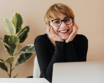 Eine lächelnde Frau mit kurzen Haaren und Brille sitzt an einem Schreibtisch, ihre Hände stützen ihr Gesicht. Vor ihr steht ein Laptop, daneben ein Smartphone und ein Stift. Im Hintergrund ist eine Pflanze sichtbar.