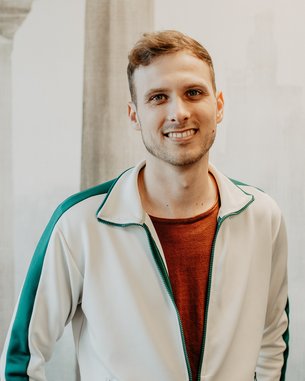 Young man with short hair, smiling, wearing a white jacket with green accents and a red T-shirt. The background is neutral.