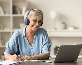 An older woman with headphones smiles as she works on a laptop and takes notes.