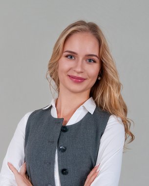 Eine Frau mit langen, blonden Haaren trägt eine graue Weste über einem weißen Hemd und steht mit verschränkten Armen vor einem neutralen Hintergrund.