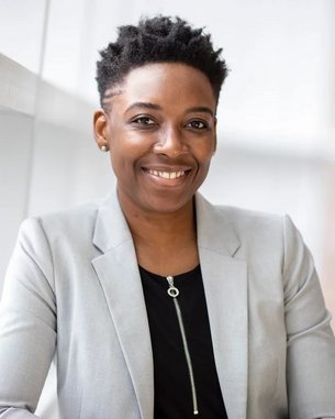A smiling woman with short, curly hair is wearing a gray blazer over a black top. She is sitting at a table and posing in a modern, bright room with large windows.
