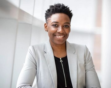 A smiling woman with short, curly hair is wearing a gray blazer over a black top. She is sitting at a table and posing in a modern, bright room with large windows.