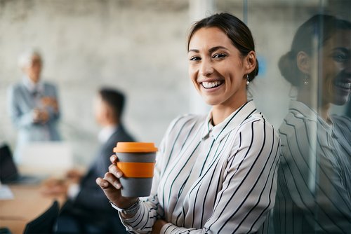 Eine lächelnde Frau mit gestreiftem Hemd hält einen Kaffeebecher in der Hand. Im Hintergrund sind zwei Personen in einem Besprechungsraum zu sehen. Die Atmosphäre wirkt professionell und modern.